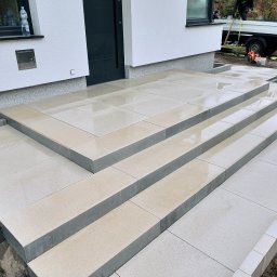 Bruk-Mat - Nowe, jasne, prostokątne płyty chodnikowe tworzące schody wejściowe do domu, z widocznymi śladami wilgoci po deszczu, w tle biała elewacja i fragment samochodu dostawczego z narzędziami.
