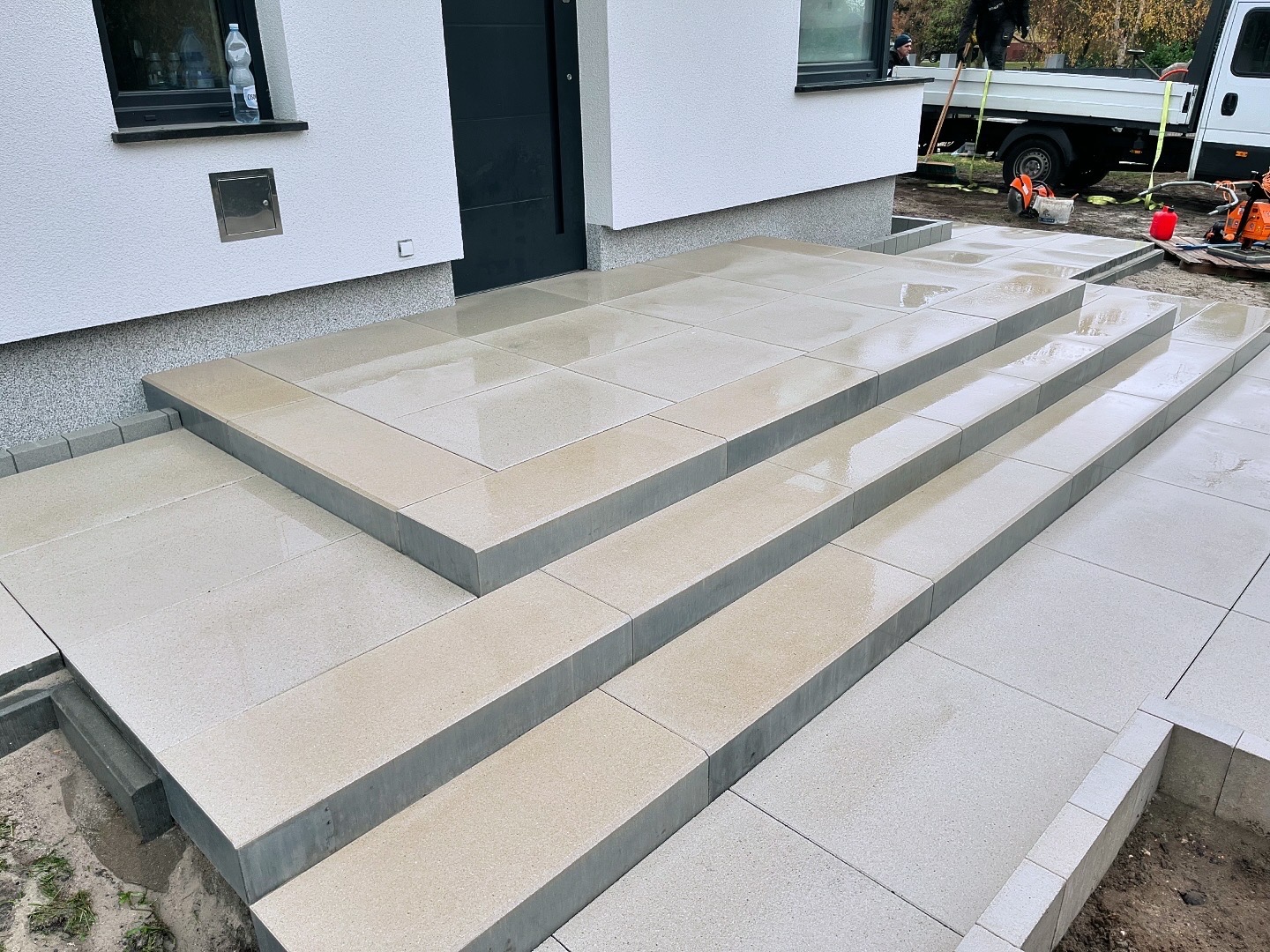 Nowe, jasne, prostokątne płyty chodnikowe tworzące schody wejściowe do domu, z widocznymi śladami wilgoci po deszczu, w tle biała elewacja i fragment samochodu dostawczego z narzędziami.