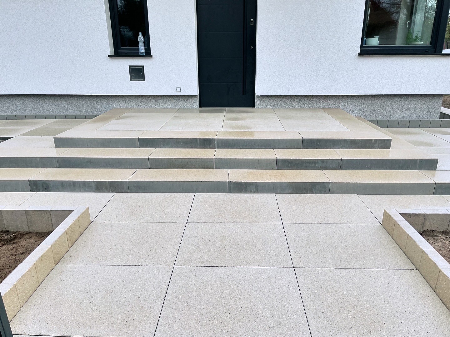 Nowoczesne wejście do domu z jasnych, kwadratowych płyt betonowych, prowadzące po schodach do ciemnych drzwi wejściowych. Widoczne dwa małe okna po bokach drzwi. Minimalistyczny design.