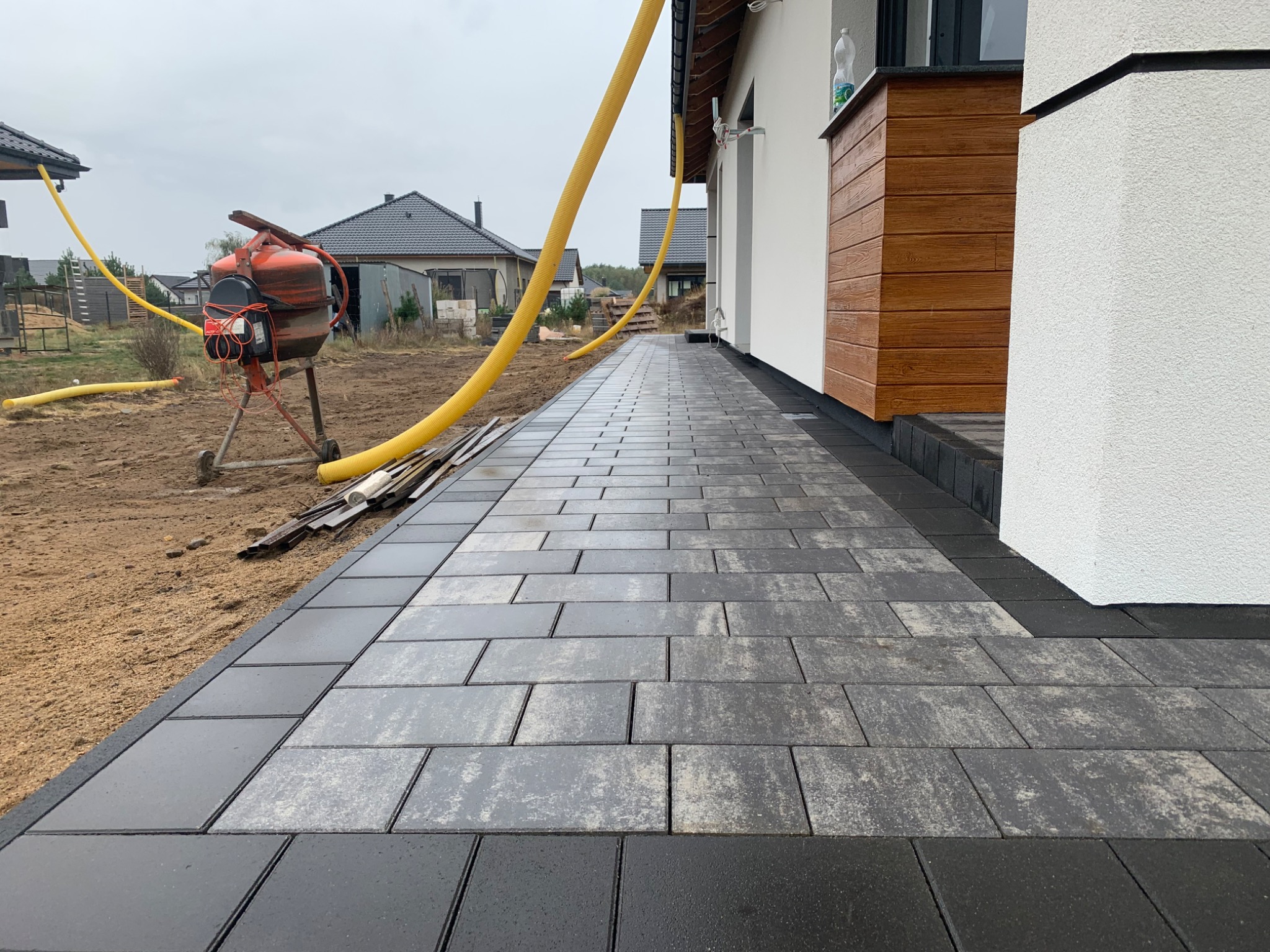 Świeżo ułożona, wilgotna kostka brukowa w odcieniach szarości i czerni prowadząca do domu z elewacją w bieli i drewnie, widoczna betoniarka i żółte rury drenażowe w tle.
