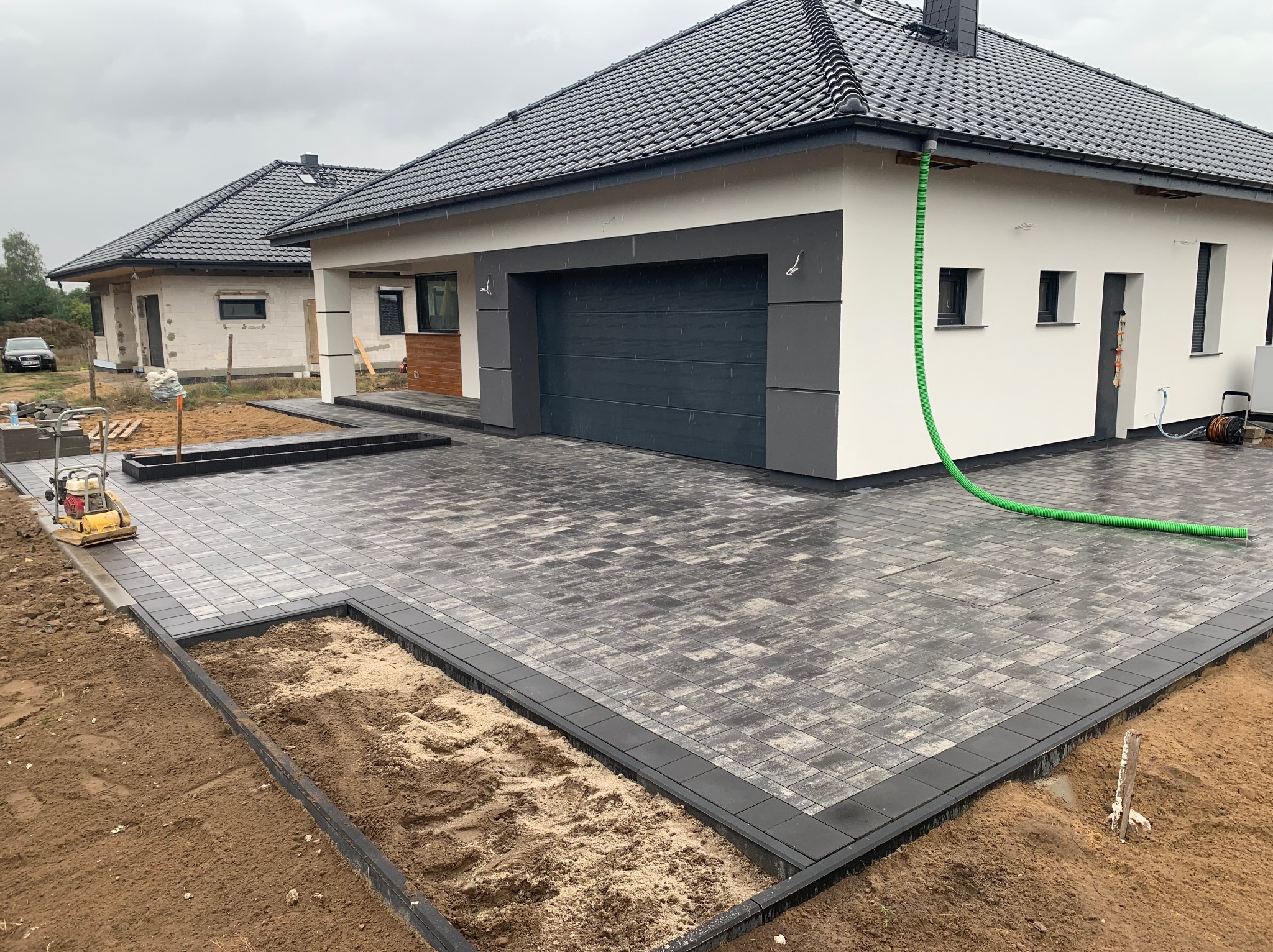 Ułożona szara kostka brukowa na podjeździe nowego domu, z widoczną zagęszczarką i fragmentami terenu przygotowanego pod dalsze prace wykończeniowe.