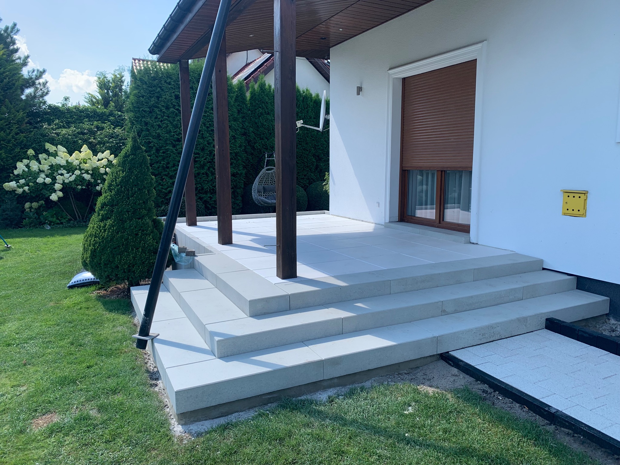 Nowy taras z jasnoszarych płyt, schody prowadzące do domu z brązowymi roletami, widoczny fragment ogrodu z zielonym trawnikiem i krzewami.