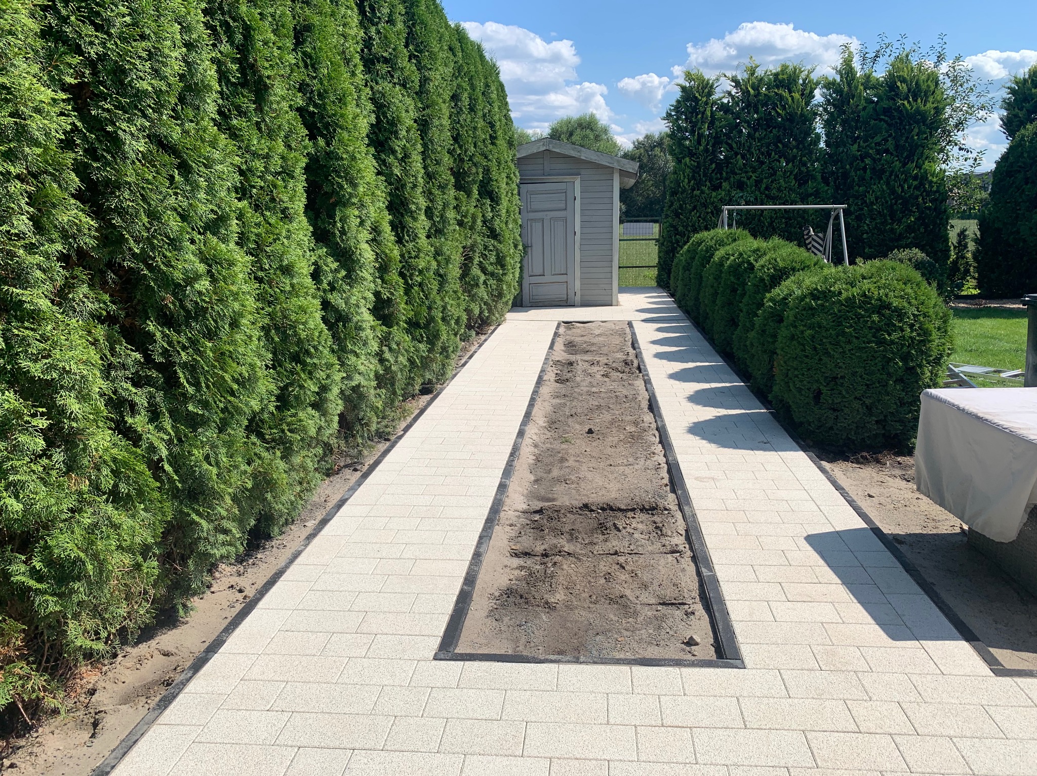 Jasna kostka brukowa ułożona wzdłuż ścieżki ogrodowej z wykopem na drenaż liniowy, obok szpaler z tuj i mały drewniany budynek gospodarczy w tle.