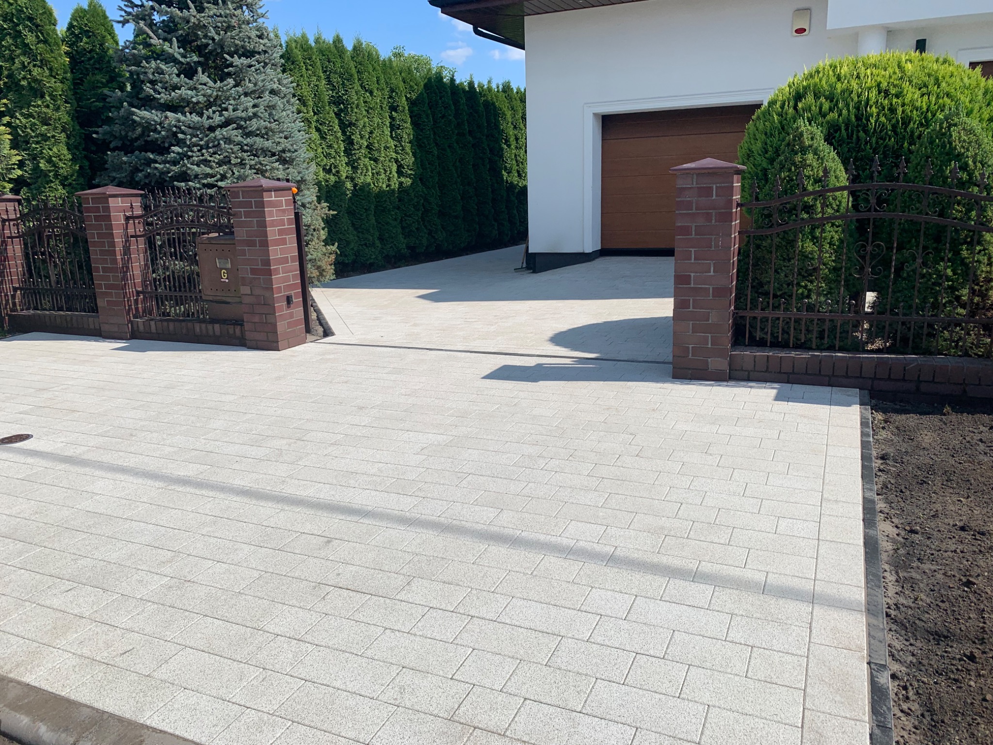 Jasnoszara kostka brukowa ułożona na podjeździe do garażu, widoczna brama z ceglanymi słupkami i metalowym ogrodzeniem, w tle zadbane tuje i biały dom.