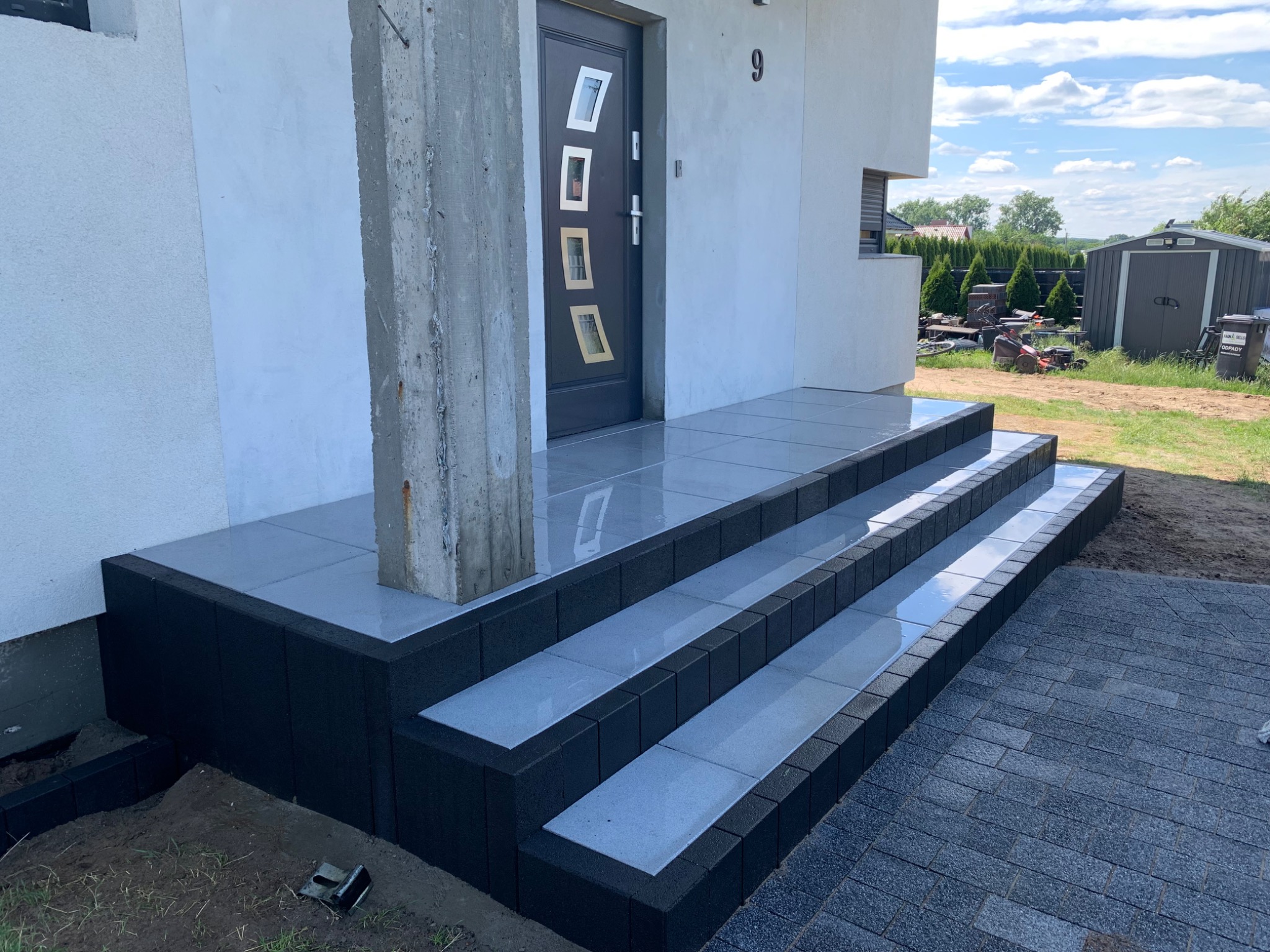 Nowe schody wejściowe wyłożone szarymi płytkami i czarnym obrzeżem, prowadzące do nowoczesnego domu z numerem 9, z fragmentem nowo ułożonej kostki brukowej przed schodami.