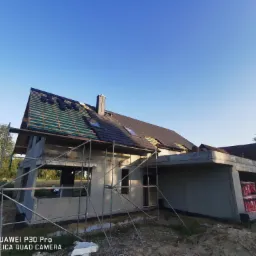 Budowa domu jednorodzinnego w stanie surowym otwartym, z częściowo ułożonym pokryciem dachowym z dachówki ceramicznej, widoczne deski i membrana dachowa, rusztowanie przy ścianach.