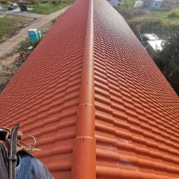 Długi, pomarańczowy dach z dachówki ceramicznej widziany z góry, z perspektywy pracownika w uprzęży, w tle krajobraz z domami i zielenią.