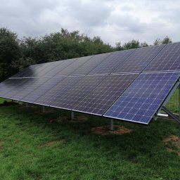 Fenix - Panel słoneczny ustawiony na metalowej konstrukcji na trawniku, widoczne drzewa i ogrodzenie w tle.