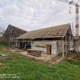 Remont dachu starego budynku gospodarczego z użyciem rusztowania i dźwigu; widoczna konstrukcja więźby dachowej i fragmenty starego pokrycia.