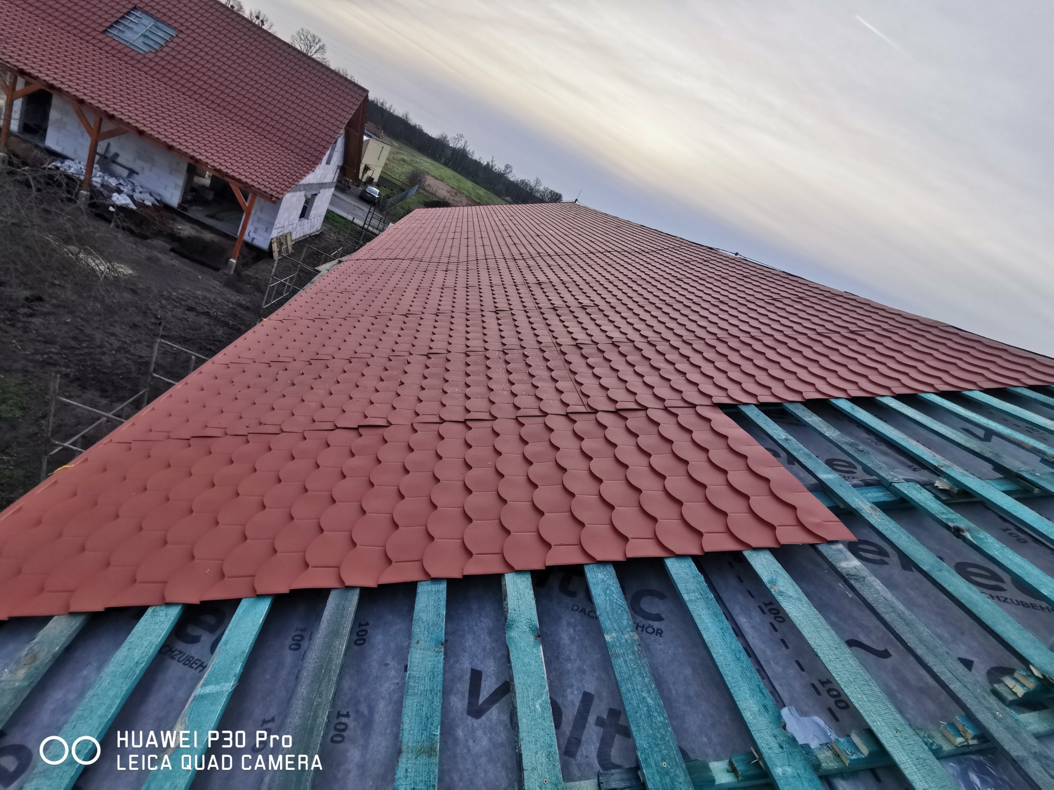 Częściowo pokryty dach z czerwonej dachówki karpiówki na deskowaniu z zielonych łat, widok z góry, w tle budynek w stanie surowym otwartym i pochmurne niebo.