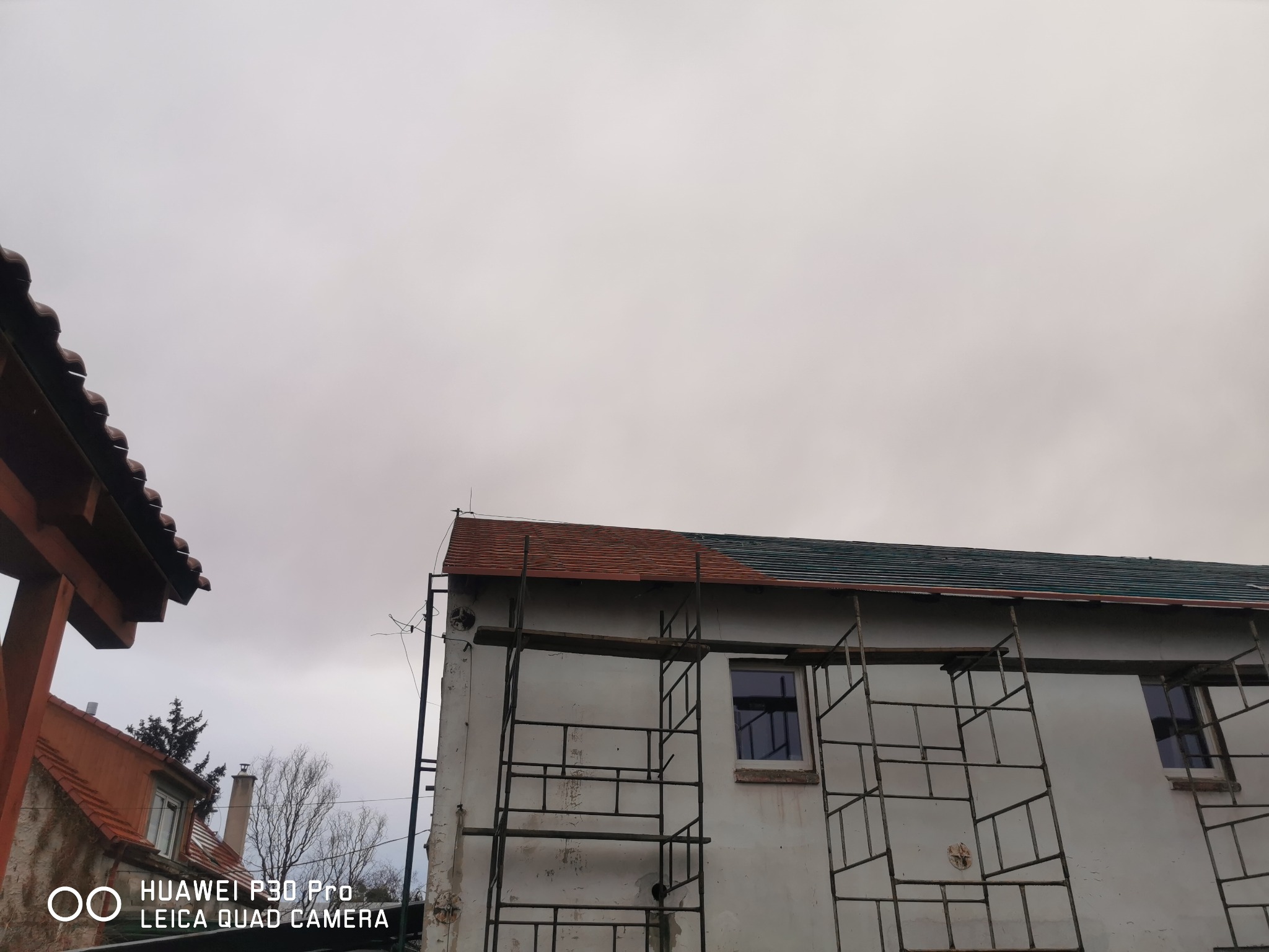 Fragment dachu z częściowo ułożoną czerwoną dachówką i ciemnozielonym pokryciem, widoczny spod rusztowania przy białej ścianie budynku w pochmurny dzień.