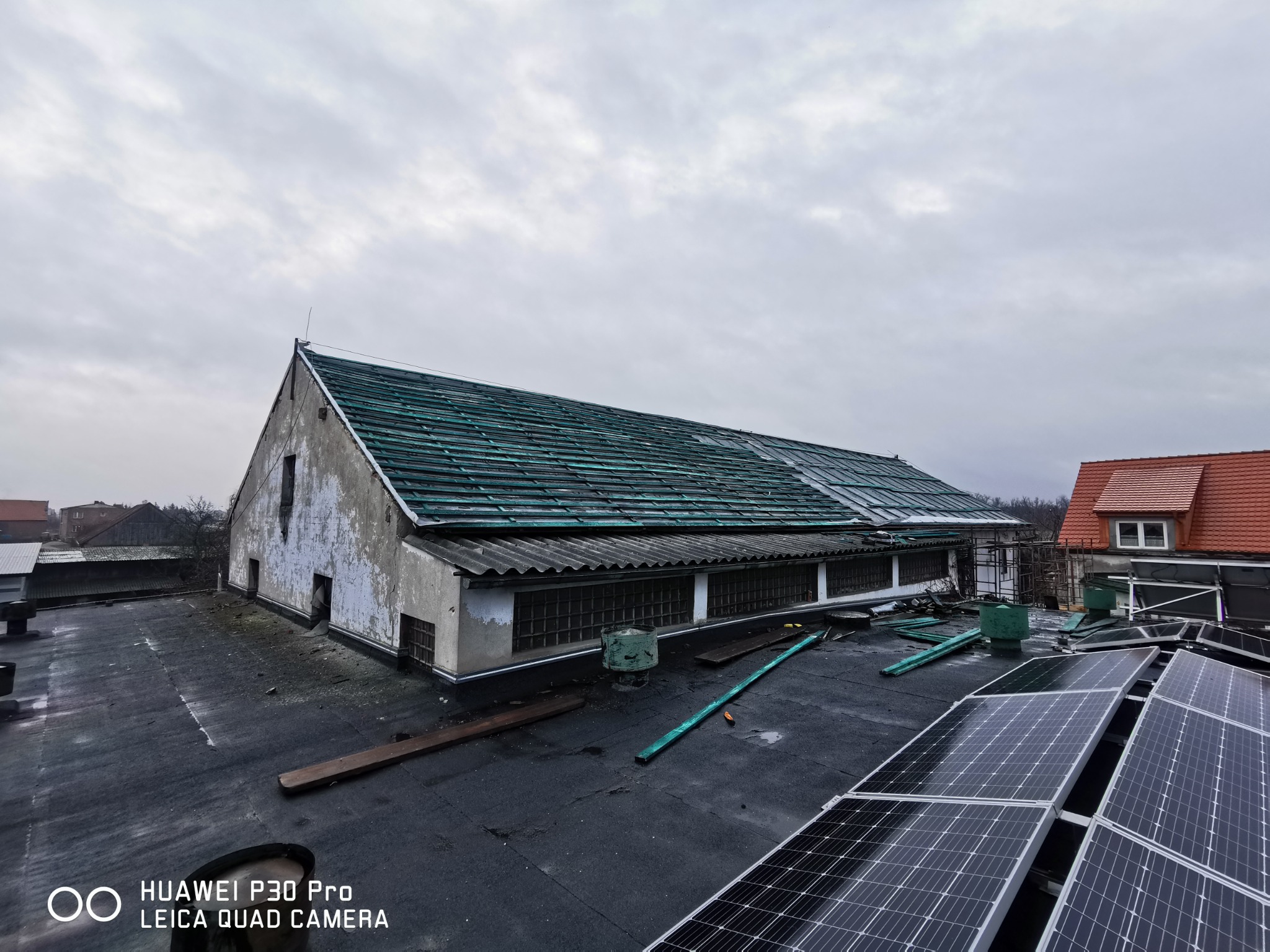 Wymiana dachu z zielonej blachy falistej na starym budynku gospodarczym w Oławie, widoczne panele fotowoltaiczne na dachu. Częściowo zdjęte poszycie i przygotowane materiały.