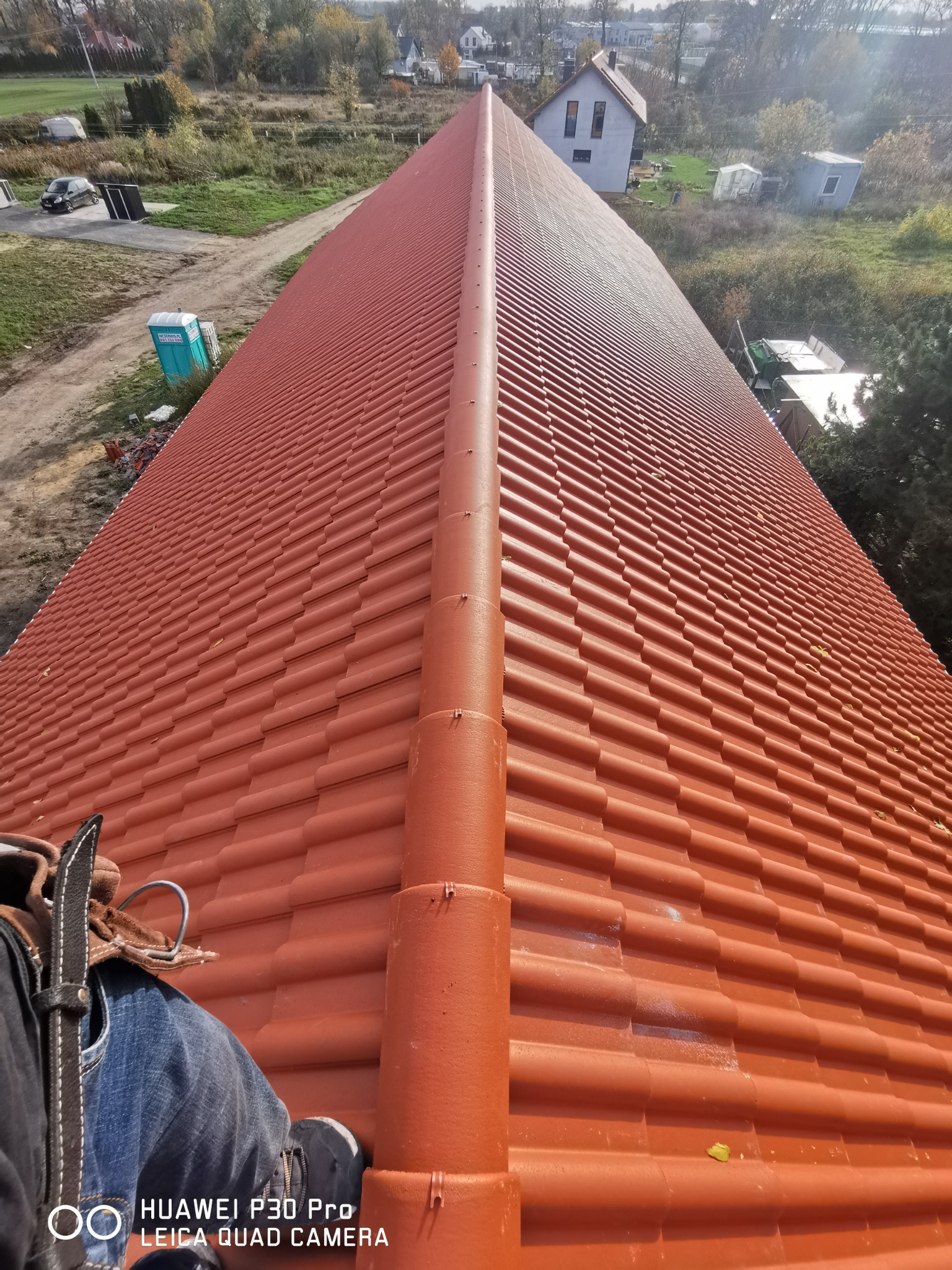 Długi, pomarańczowy dach z dachówki ceramicznej widziany z góry, z perspektywy pracownika w uprzęży, w tle krajobraz z domami i zielenią.