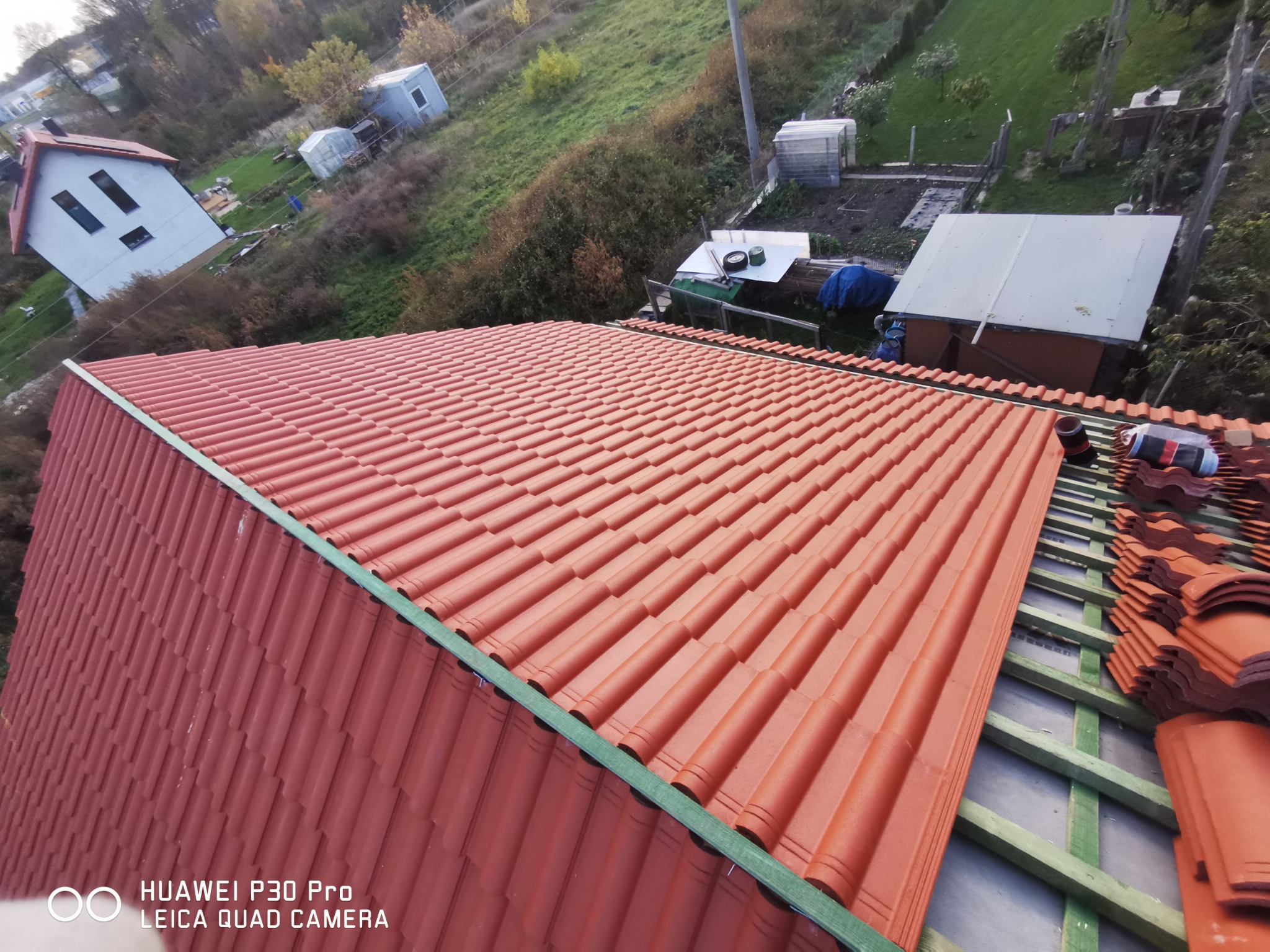 Widok z góry na fragment dachu z czerwoną dachówką ceramiczną, ułożoną częściowo na zielonej konstrukcji drewnianej, w tle budynki gospodarcze i zieleń.