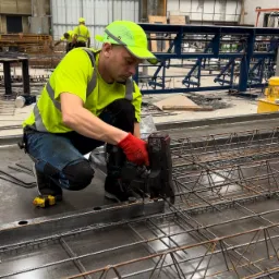 Pracownik w odblaskowej odzieży łączy zbrojenie na metalowej płycie za pomocą specjalistycznego narzędzia pneumatycznego, w tle widoczna konstrukcja stalowa i inni pracownicy.