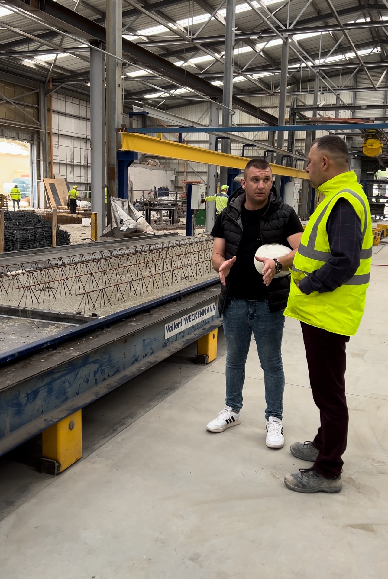 Dwóch mężczyzn rozmawia obok formy zalewanej betonem ze zbrojeniem w hali produkcyjnej, widoczny napis Vollert-Weckenmann.