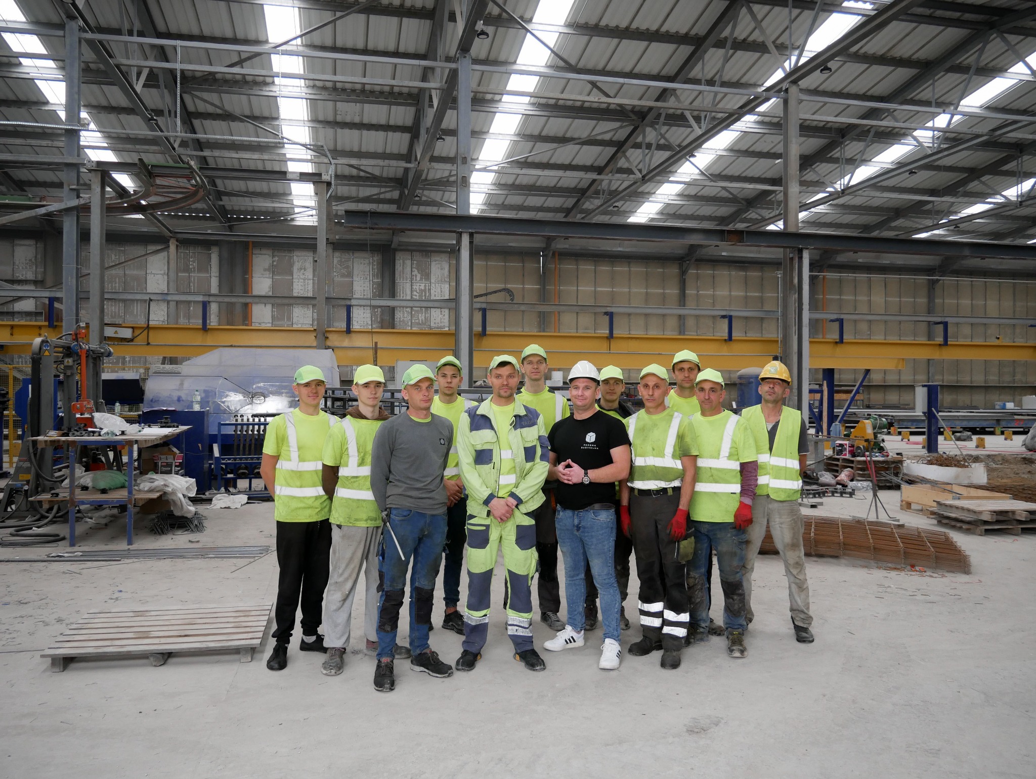 Grupa pracowników budowlanych w odblaskowych kamizelkach i czapkach, stojąca w hali produkcyjnej na tle maszyn i konstrukcji stalowych.