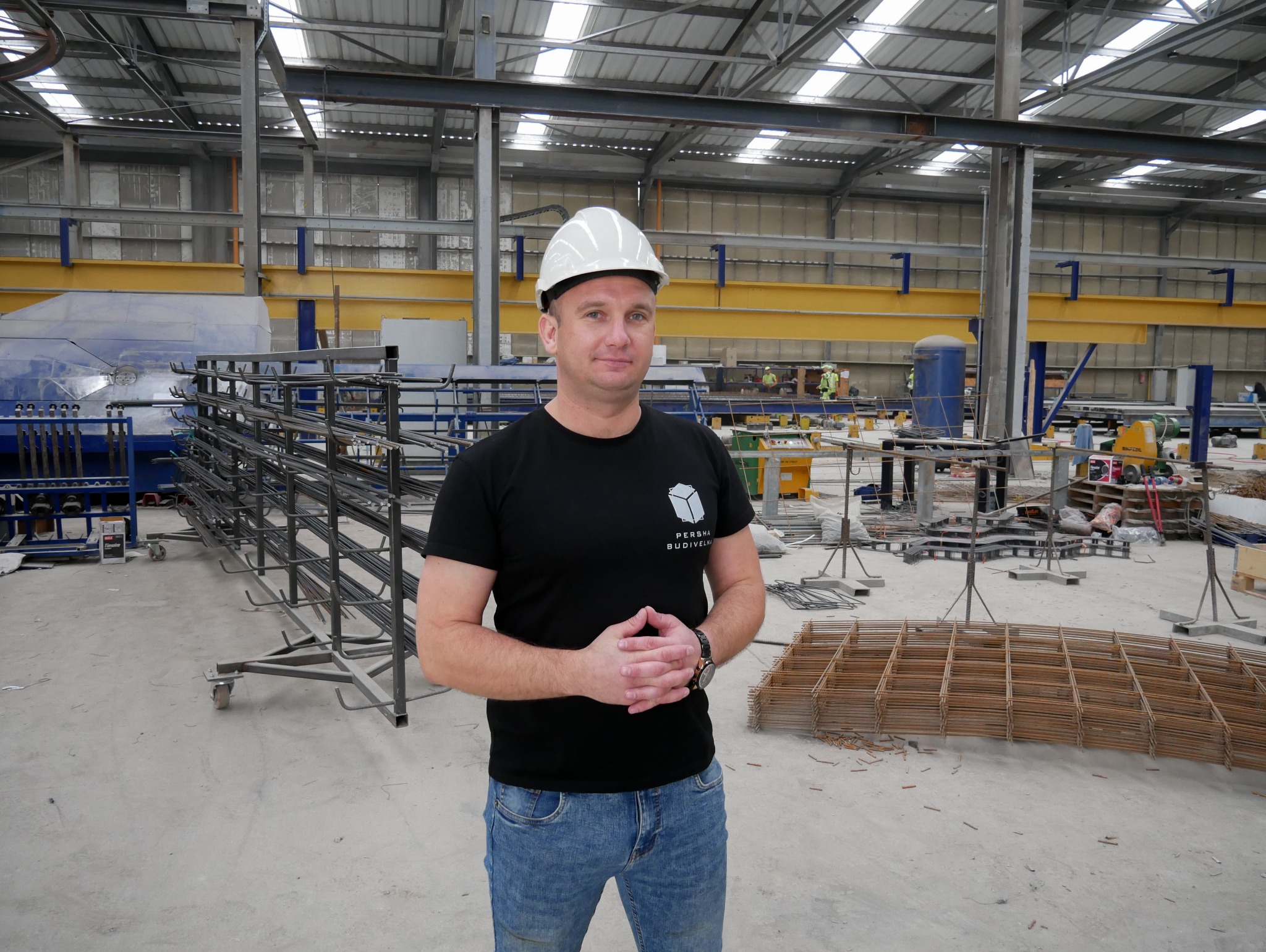 Pracownik w kasku na hali produkcyjnej z prefabrykatami zbrojeniowymi i maszyną do ich produkcji w tle.