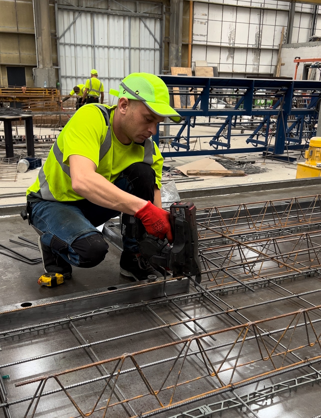 Pracownik w odblaskowej odzieży łączy zbrojenie na metalowej płycie za pomocą specjalistycznego narzędzia pneumatycznego, w tle widoczna konstrukcja stalowa i inni pracownicy.