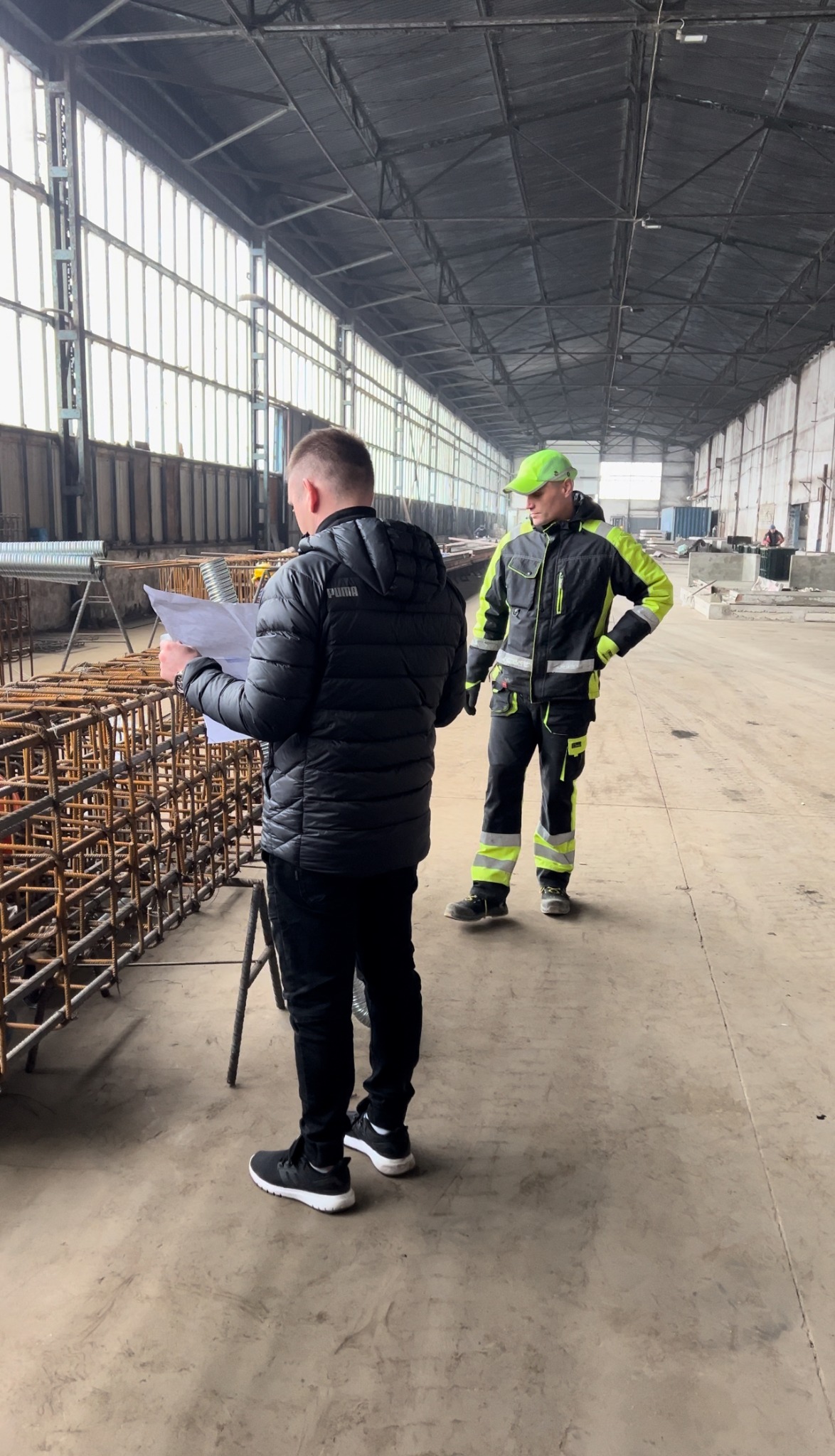 Dwóch mężczyzn w odzieży roboczej w hali produkcyjnej, jeden czyta plan przy zbrojeniu, drugi stoi obok z ręką na biodrze, w tle widoczne okna i konstrukcja dachu.