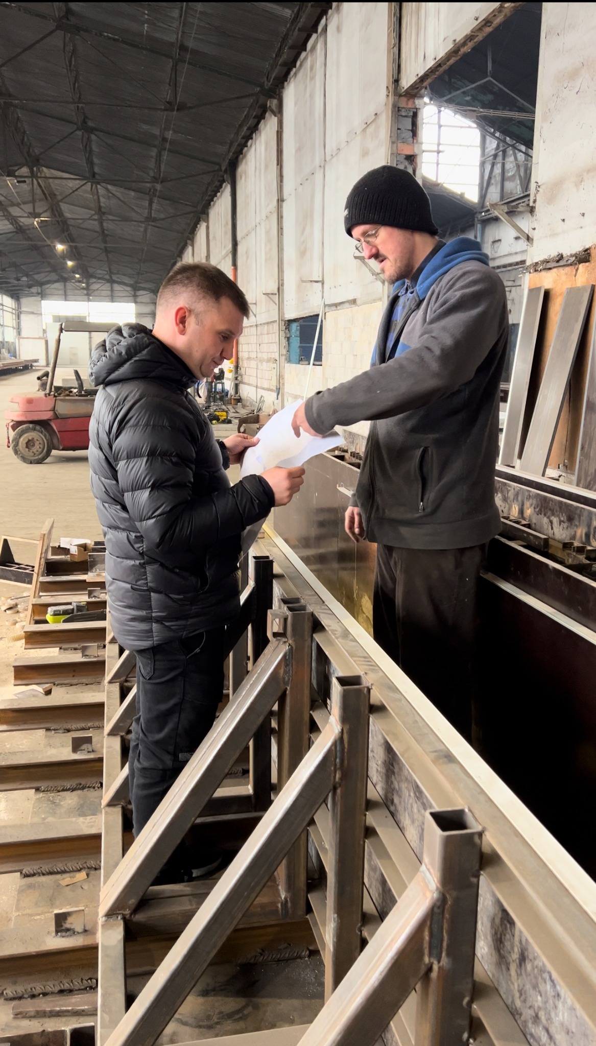 Dwóch mężczyzn w hali produkcyjnej omawia plan na papierze, stojąc obok metalowej konstrukcji z profili zamkniętych, w tle widać wózek widłowy.