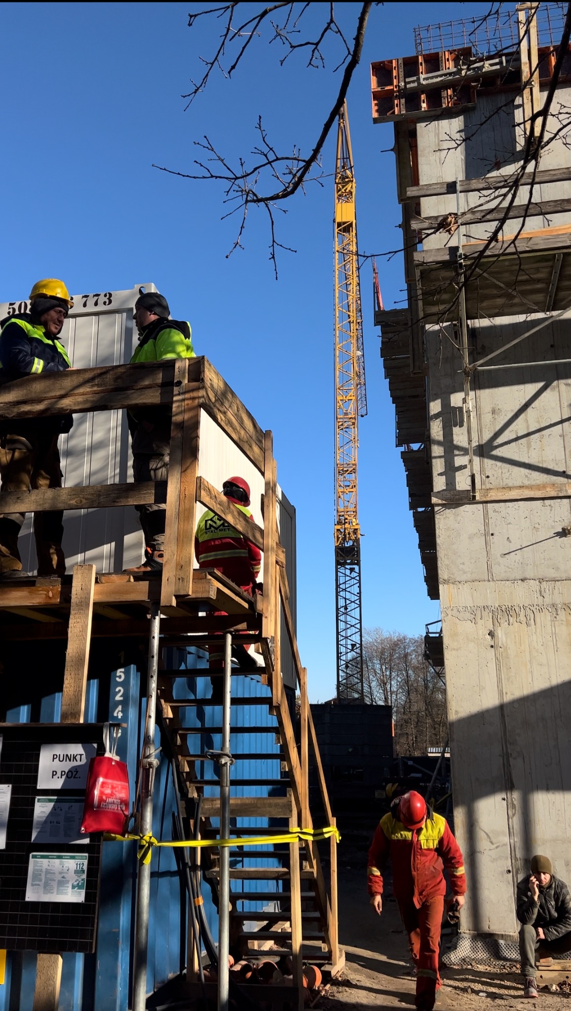 Praca zbrojarzy na budowie w Warszawie: widok na rusztowanie, żuraw i szkielet budynku w słoneczny dzień, z pracownikami w hełmach i kombinezonach.