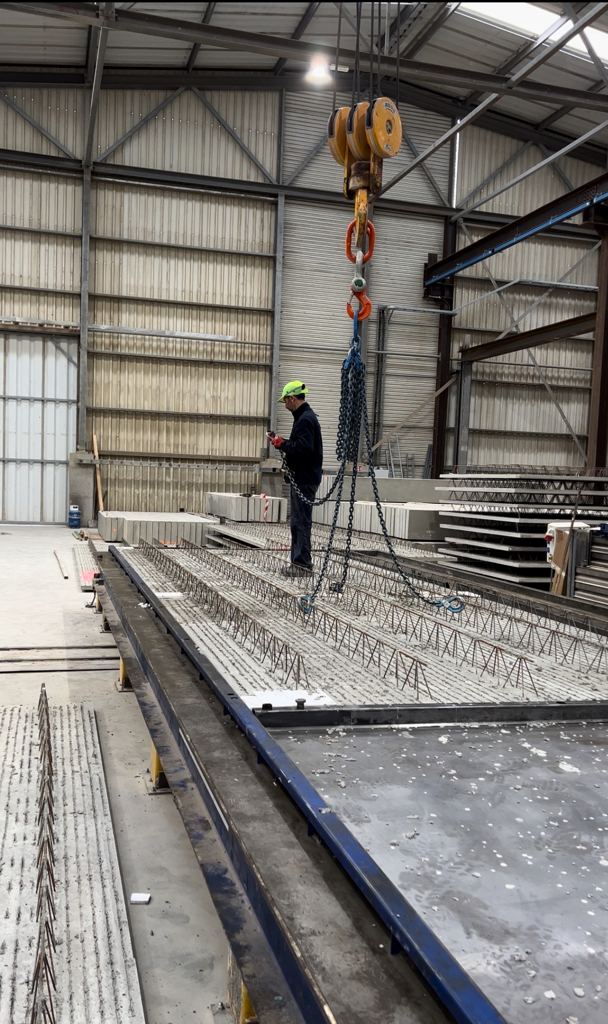 Pracownik w kasku i rękawicach, obsługujący suwnicę łańcuchową nad prefabrykowanym elementem zbrojonym na hali produkcyjnej.