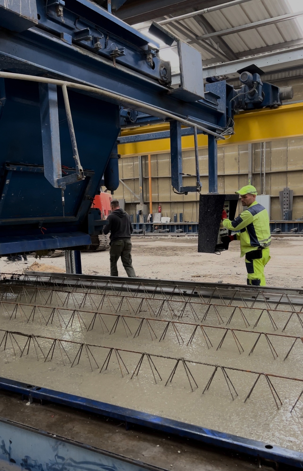 Proces zalewania betonem konstrukcji zbrojeniowej w hali produkcyjnej, pracownik w odblaskowym ubraniu obsługuje maszynę, drugi pracownik w tle.