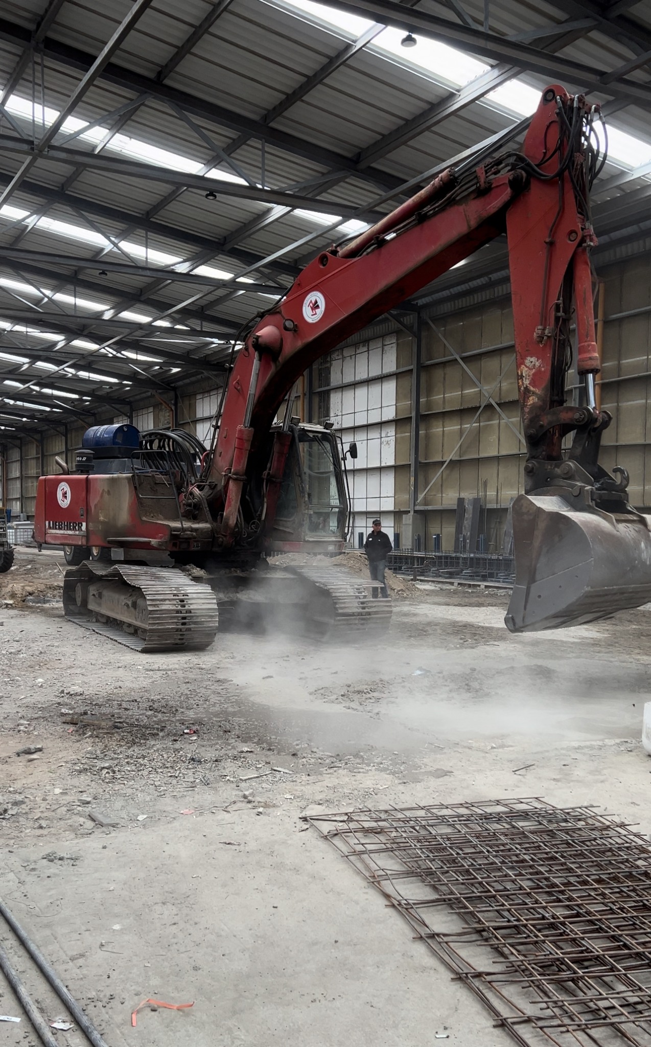 Czerwona koparka Liebherr w hali przemysłowej, operator stoi w tle, w pierwszym planie siatka zbrojeniowa.