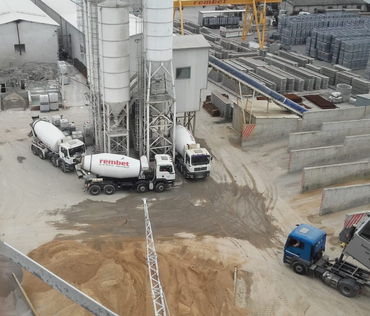 Widok z góry na plac budowy z betoniarkami, silosami, stertami prefabrykatów betonowych i suwnicą w tle.