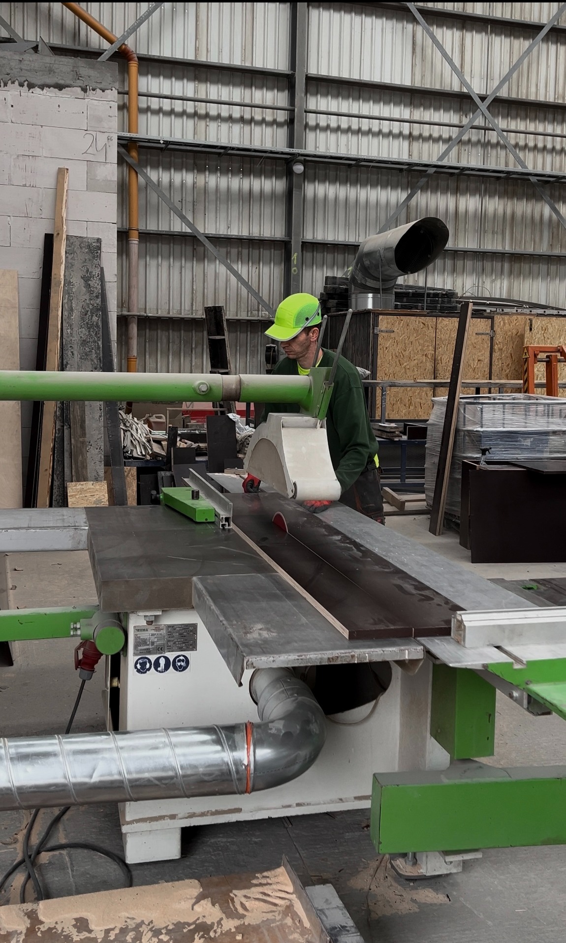 Pracownik w zielonym kasku tnie deskę na pile stołowej w hali produkcyjnej.