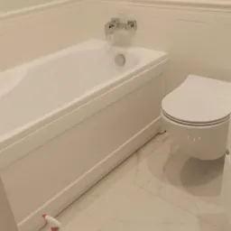 Nowoczesna, biała łazienka z wanną, podwieszaną toaletą i marmurową podłogą. Widoczny środek czyszczący obok wanny.