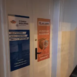 Dwa plakaty reklamowe na białych drzwiach: po lewej stronie plakat firmy Power Holding oferującej ubezpieczenia, nieruchomości i kredyty hipoteczne, a po prawej plakat Ship Center z ofertą...