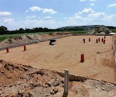 Przygotowany teren pod budowę: wyrównany piasek, wystające pionowe rury kanalizacyjne, w oddali hałda ziemi i zieleń.