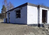 Surowy stan budynku w trakcie budowy, widoczne ściany z białych bloczków, betonowa wylewka fundamentowa, niezabudowane otwory okienne i drzwiowe, teren wokół niezagospodarowany.