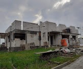 Surowy stan budynku jednorodzinnego w trakcie budowy, widoczne ściany z bloczków betonowych, częściowo zbrojone nadproża, teren wokół z fragmentami trawy i nieba.