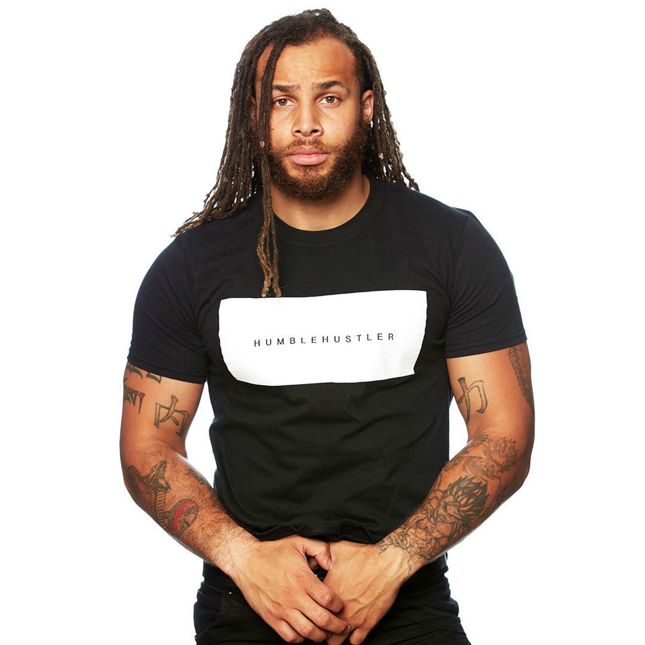 Mężczyzna z dredami i tatuażami na ramionach, ubrany w czarną koszulkę z białym prostokątem i napisem 'HUMBLEHUSTLER'.