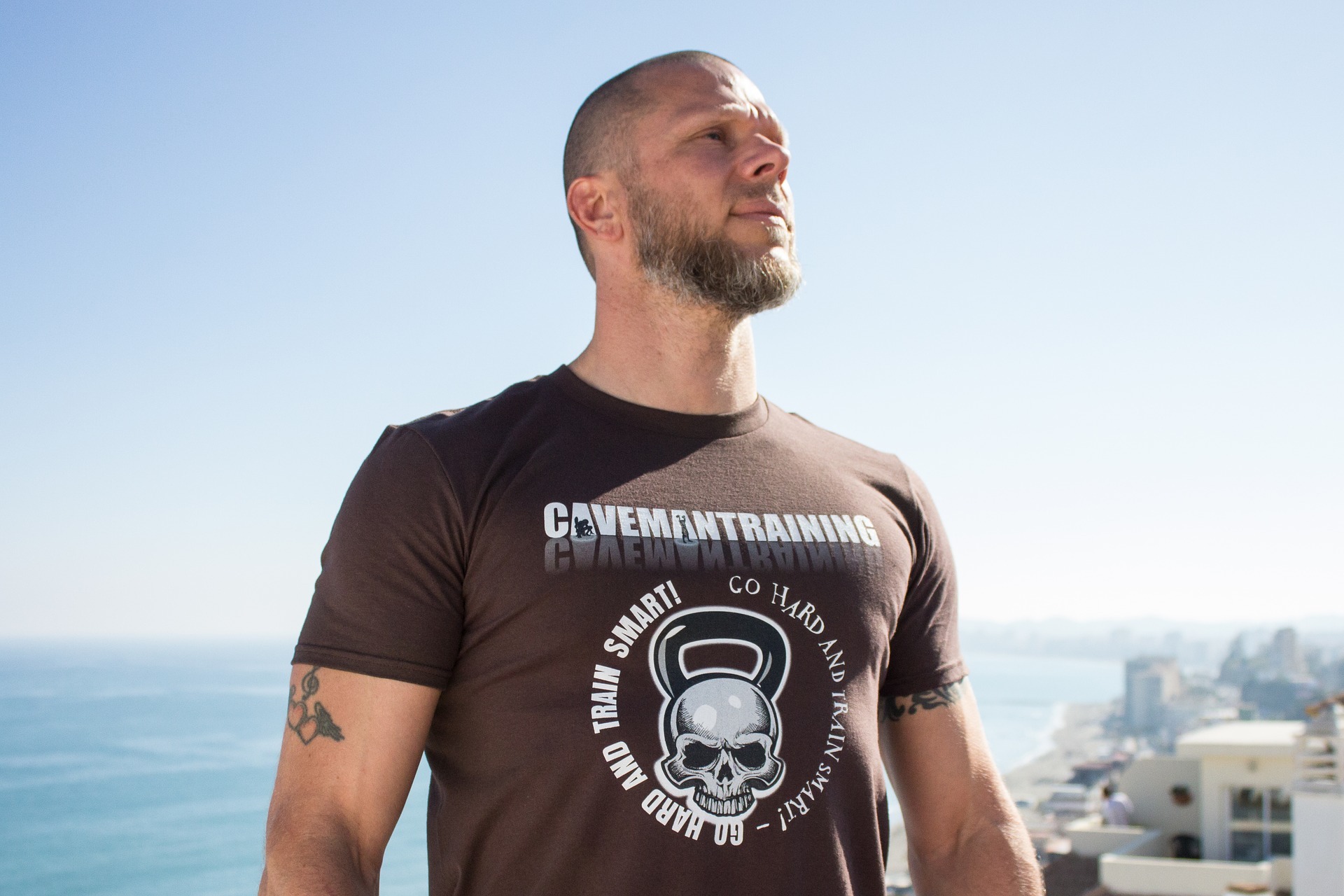 Mężczyzna z brodą w brązowej koszulce z nadrukiem 'Cavemantraining' i motywem czaszki z kettlebell, pozujący na tle panoramy Warszawy z widokiem na miasto i błękitne niebo.