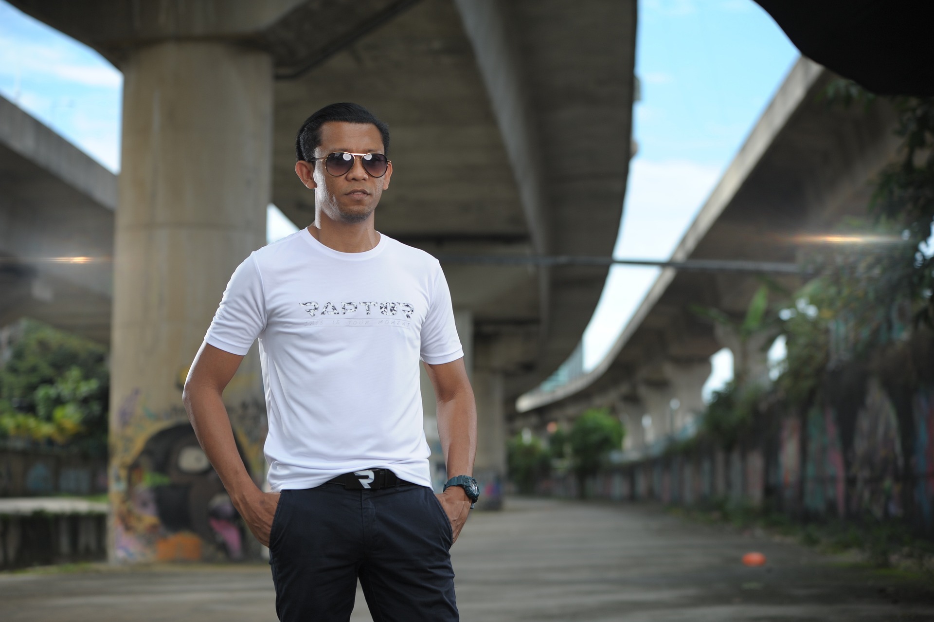 Mężczyzna w okularach przeciwsłonecznych pozuje w białej koszulce z nadrukiem i ciemnych spodniach na tle betonowego wiaduktu.