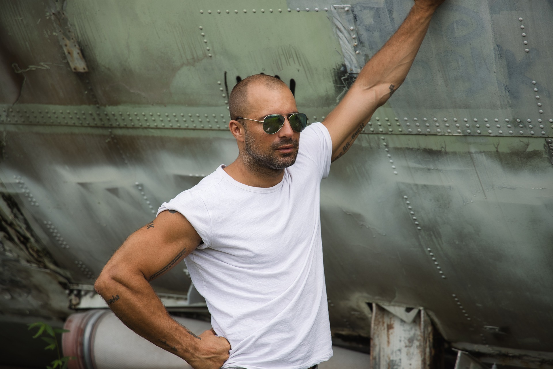 Mężczyzna w białym T-shircie i okularach przeciwsłonecznych pozuje oparty o starą, metalową konstrukcję z nitami, eksponując tatuaże na ramionach.