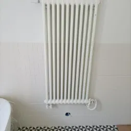 Biały, żeliwny grzejnik żeberkowy zamontowany na ścianie w łazience w Dąbrowie Górniczej, z widocznymi rurami doprowadzającymi i wzorzystą podłogą w czarno-białe trójkąty.