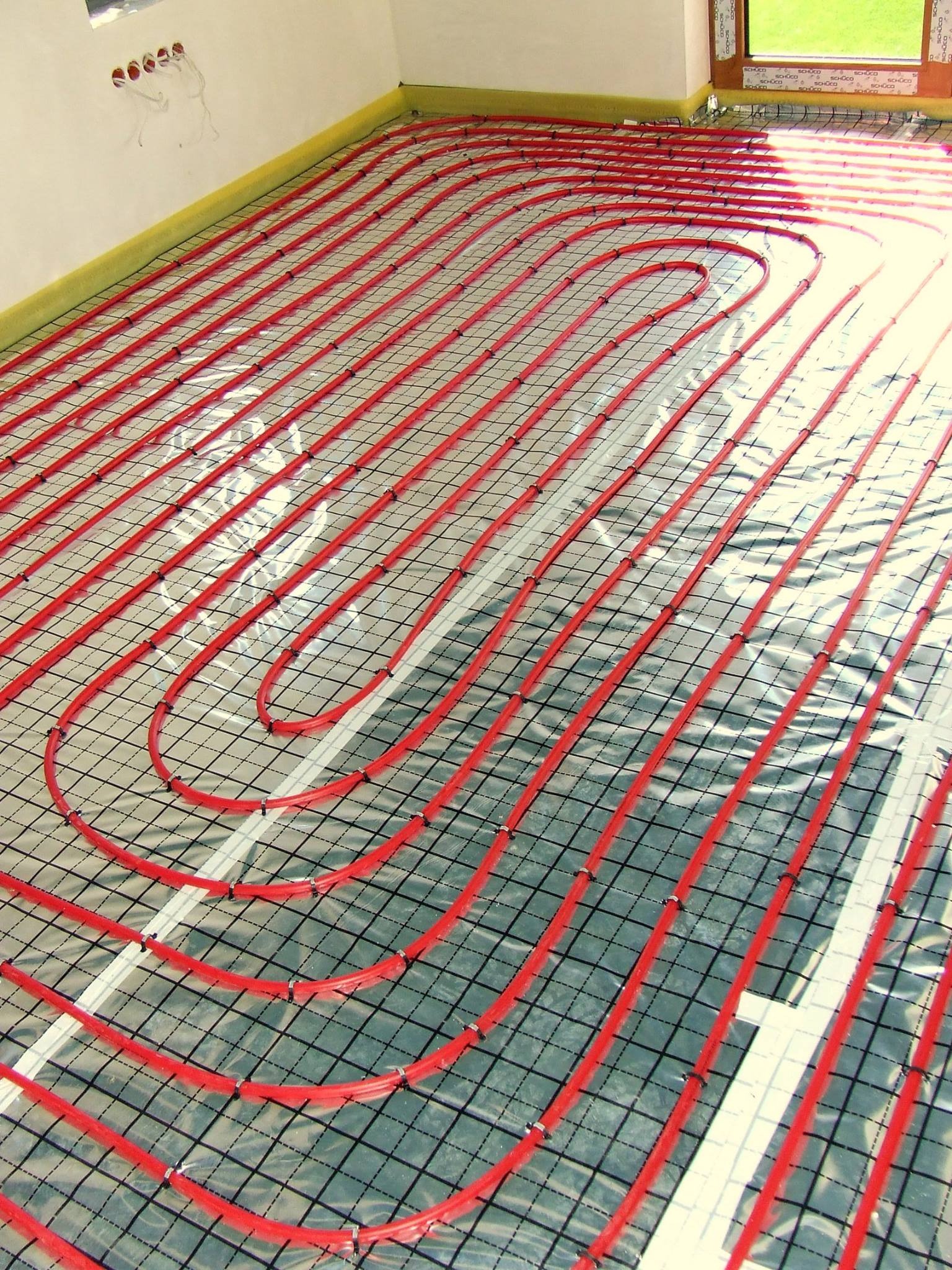 Czerwone rurki ogrzewania podłogowego ułożone na izolacji z siatką w jasnym pomieszczeniu z oknem, widoczne otwory na gniazdka elektryczne w ścianie.