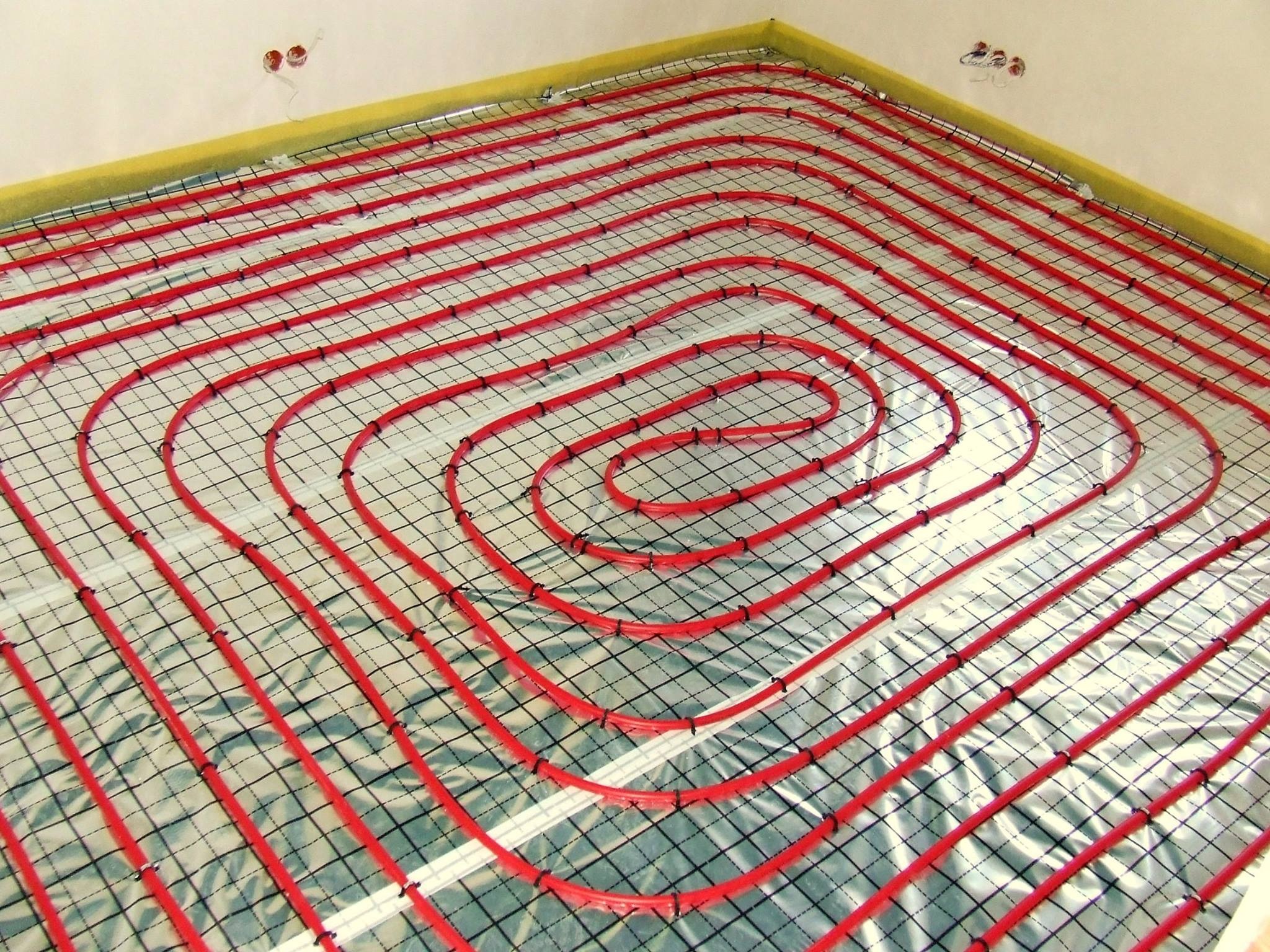 Czerwone rurki ogrzewania podłogowego ułożone spiralnie na folii izolacyjnej z czarną siatką, widoczne kable elektryczne wystające ze ściany.