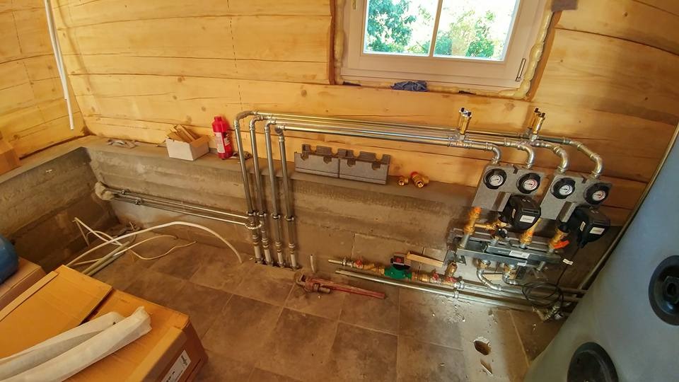 Wnętrze drewnianego domu z widoczną instalacją grzewczą: rury, pompy obiegowe, zawory, manometry, oraz narzędzia (klucz nastawny) na podłodze z płytek. Częściowo rozpakowane elementy instalacji...