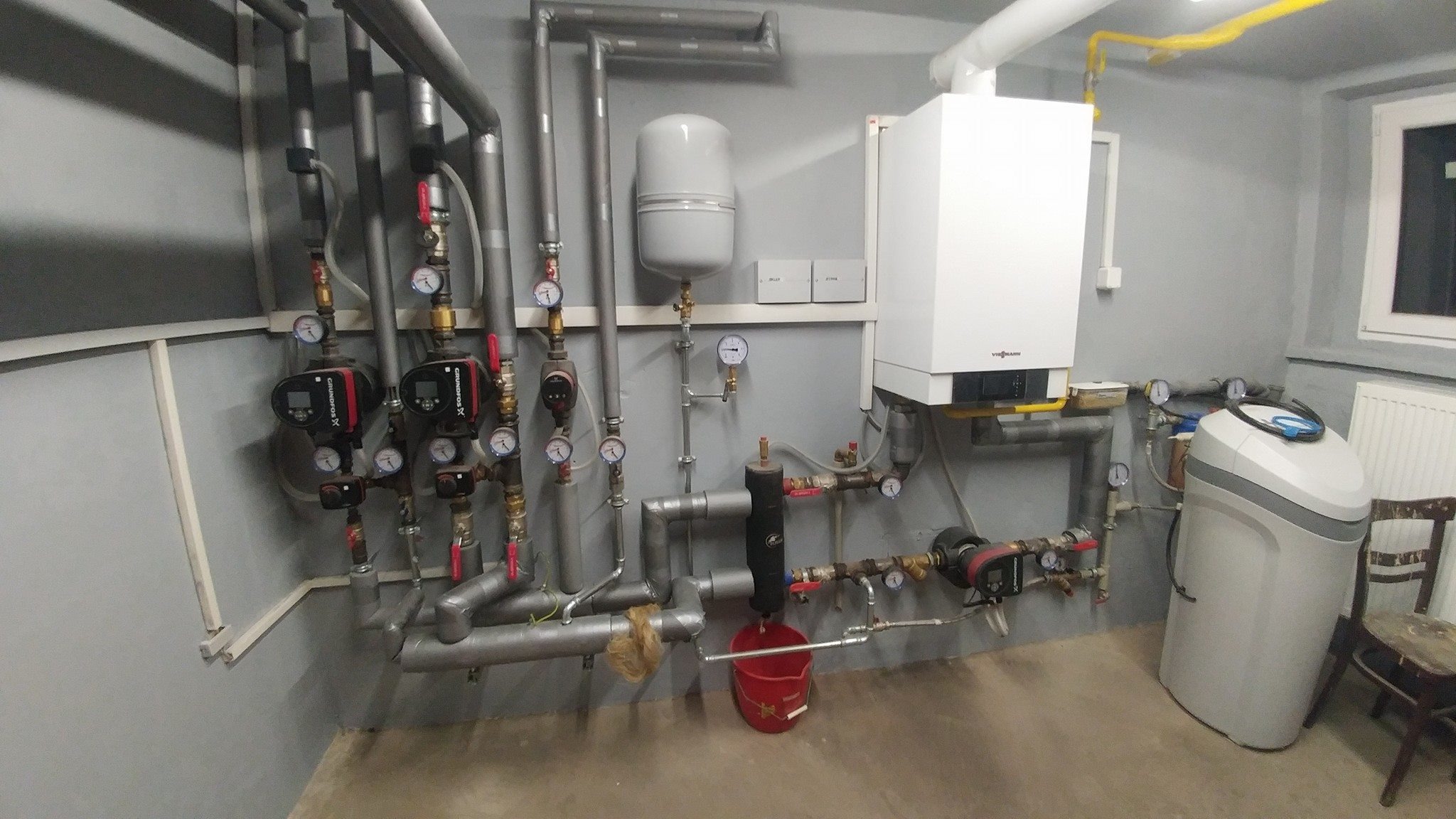 Węzeł cieplny w kotłowni z piecem gazowym Viessmann, zbiornikiem hydroforowym, pompami obiegowymi Grundfos i zmiękczaczem wody, rury w izolacji, manometry, czerwone zawory, ściany w kolorze szarym.