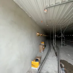 Wnętrze hali w trakcie prac budowlanych, świeżo otynkowana ściana z widocznymi śladami po szpachlowaniu, na podłodze kartony i elementy konstrukcyjne, oświetlenie sufitowe.