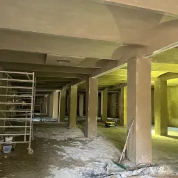 Wnętrze hali w trakcie prac wykończeniowych; widoczne rusztowania, kolumny i ściany po nałożeniu tynku, podłoga zabezpieczona folią, żółte oświetlenie.