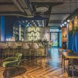 Nowoczesne wnętrze biurowe z parkietem w jodełkę, barem z podświetlanym napisem i krzesłami barowymi oraz niebieską ścianą z dekoracyjnymi elementami. Widoczne stoły z krzesłami i rośliny doniczkowe.