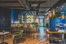 Nowoczesne wnętrze biurowe z parkietem w jodełkę, barem z podświetlanym napisem i krzesłami barowymi oraz niebieską ścianą z dekoracyjnymi elementami. Widoczne stoły z krzesłami i rośliny doniczkowe.