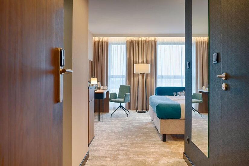 Widok z korytarza na wykończony pokój hotelowy w Warszawie z wykładziną podłogową, biurkiem, krzesłami i łóżkiem z turkusowymi akcentami.